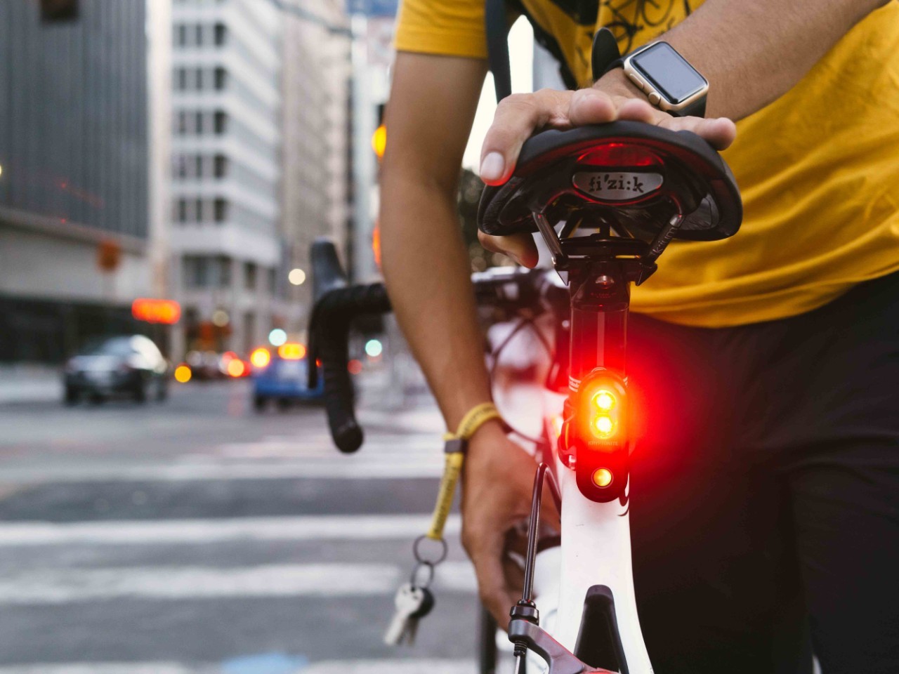 A person in a yellow shirt standing with a bicycle, with a red light illuminated under the bike seat, in an urban setting.