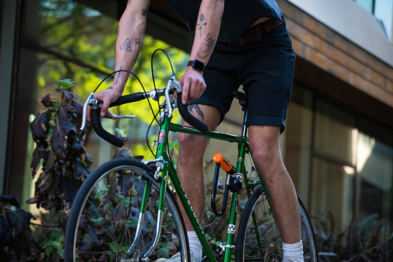 A person holding a yellow Kryptonite lock next to a pink bicycle, wearing a black shirt and a coiled keychain on their wrist.